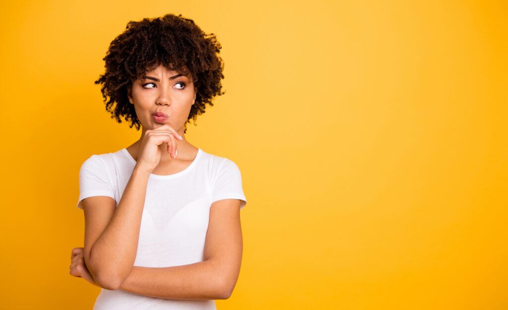 A highly sensitive person (woman) looking confused holding her hand to her chin to think, in front of a yellow wall