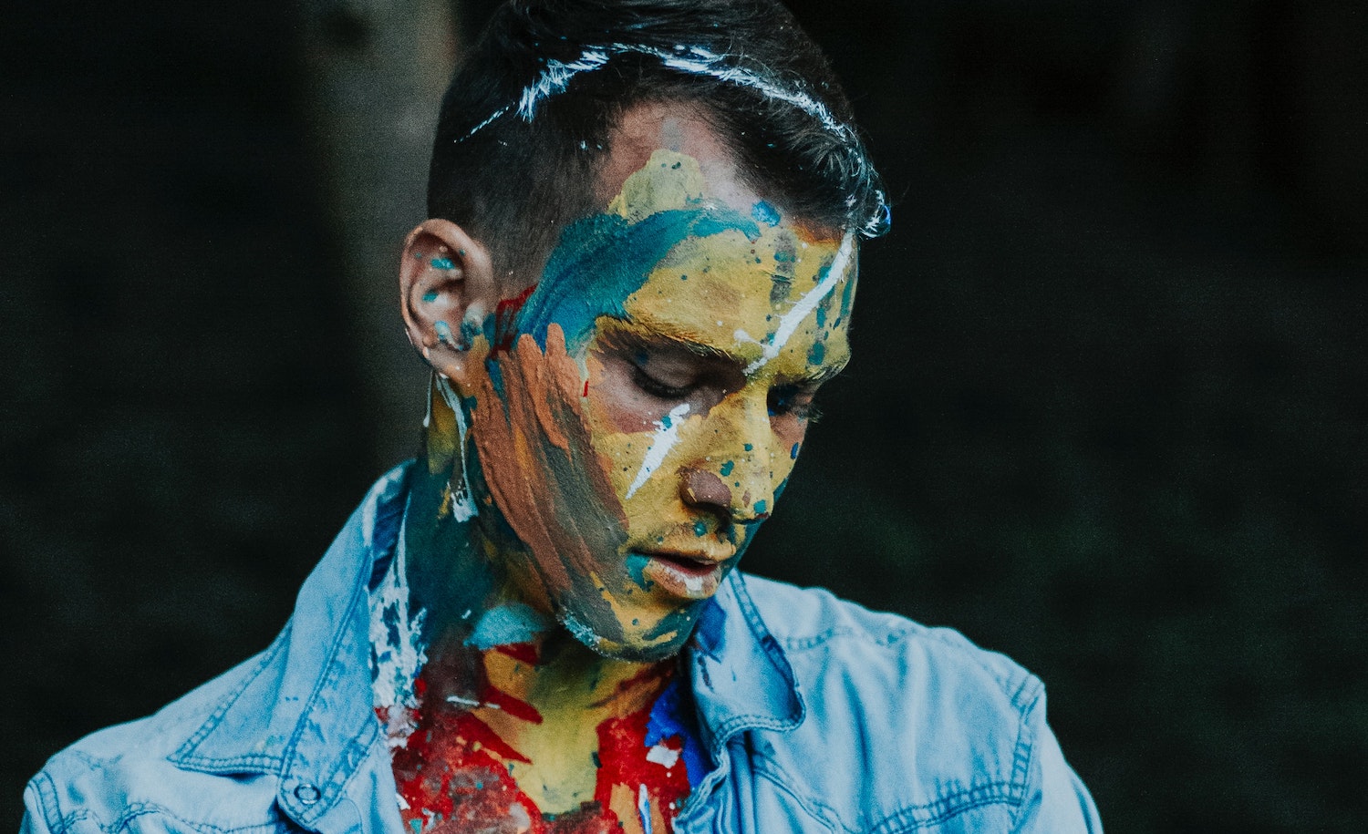 A sensitive man with paint on his face looking down in deep thought, with a dark background